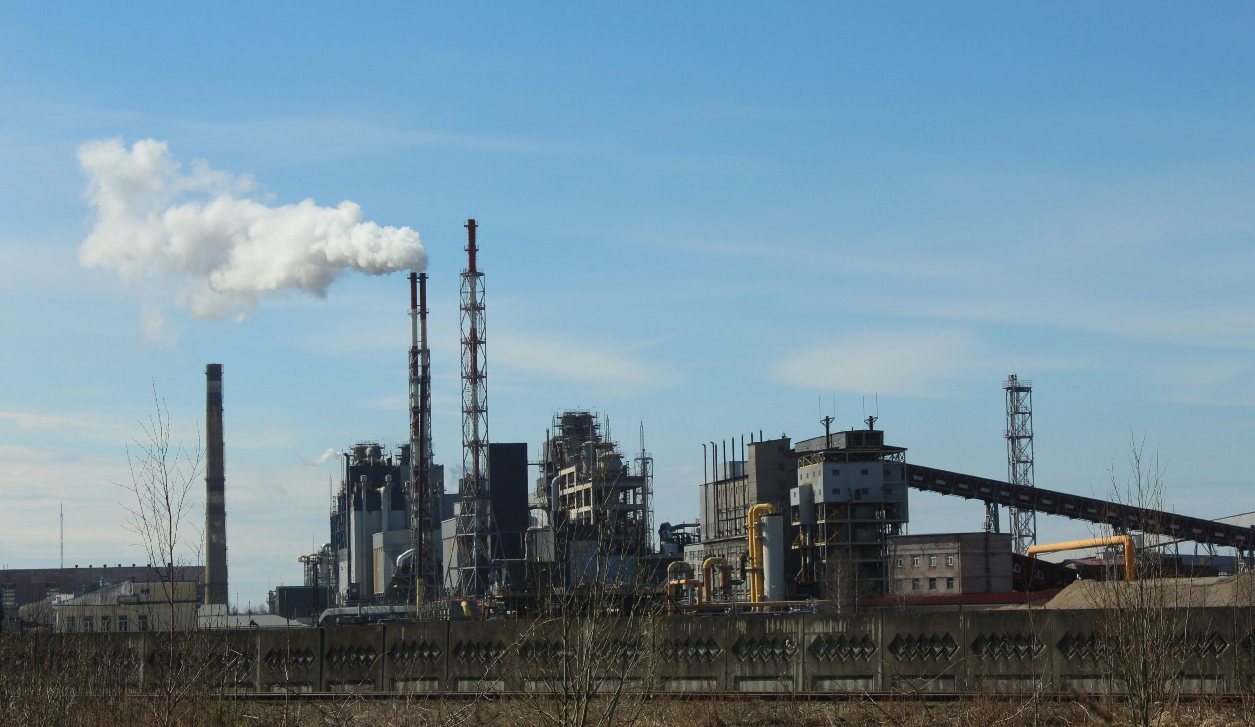 Noble Grupo Industrial Photo capturing an industrial factory with smokestacks emitting smoke under a clear blue sky.