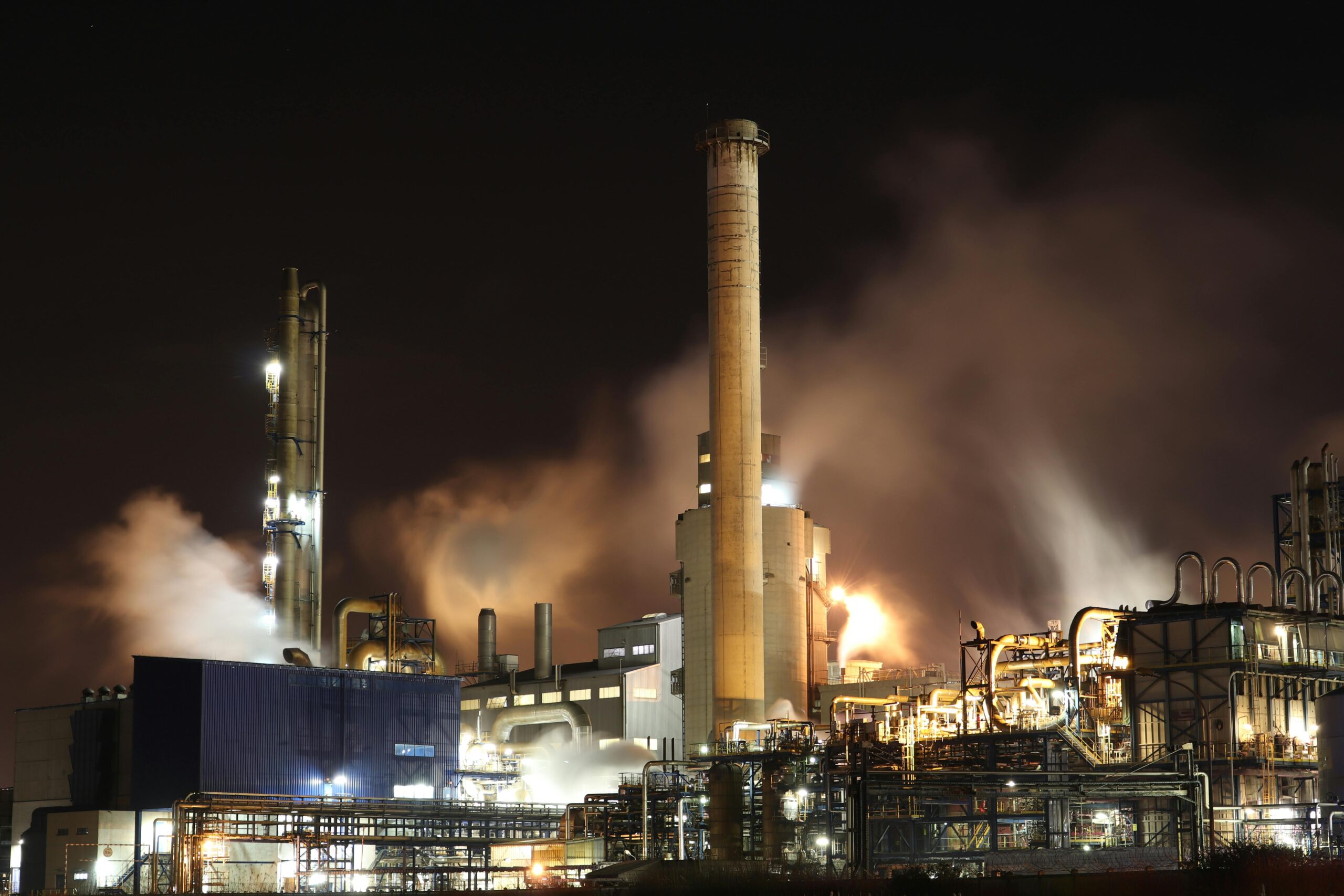 Noble Grupo Industrial A nighttime view of an industrial factory with smokestacks emitting smoke, highlighting energy production and pollution.